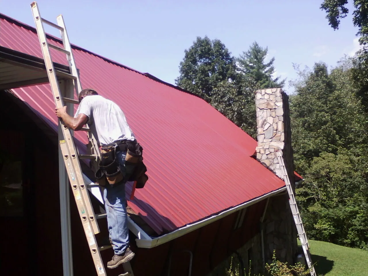 Roofer on a ladder beside a red metal roof and stone chimney — Soffit & Fascia Repair by Roofing Brothers in Orion